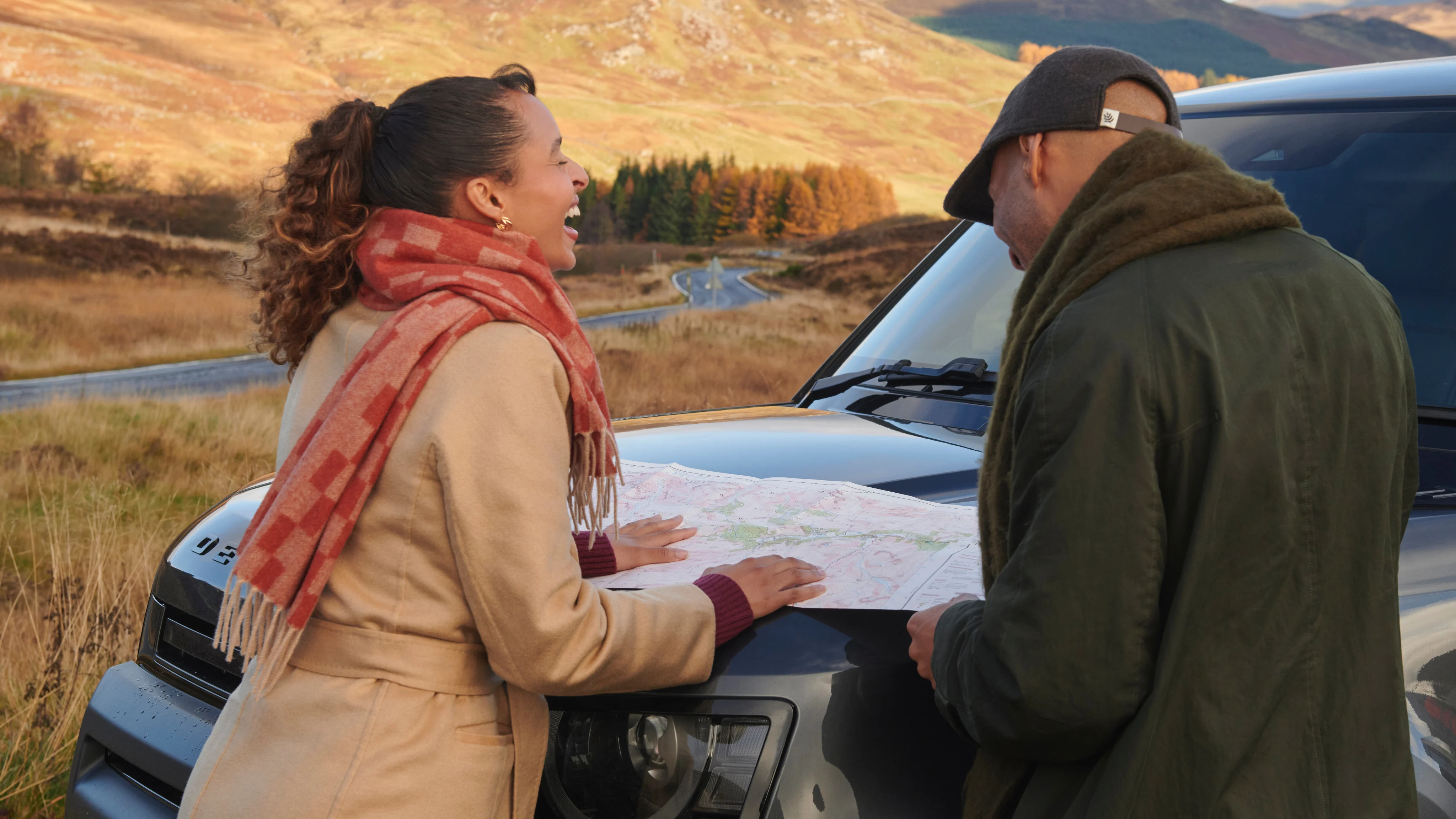 A couple consulting a map outdoors in the Scottish Highlands while planning a Highland distillery tour route, with moorland and hills stretching behind them
