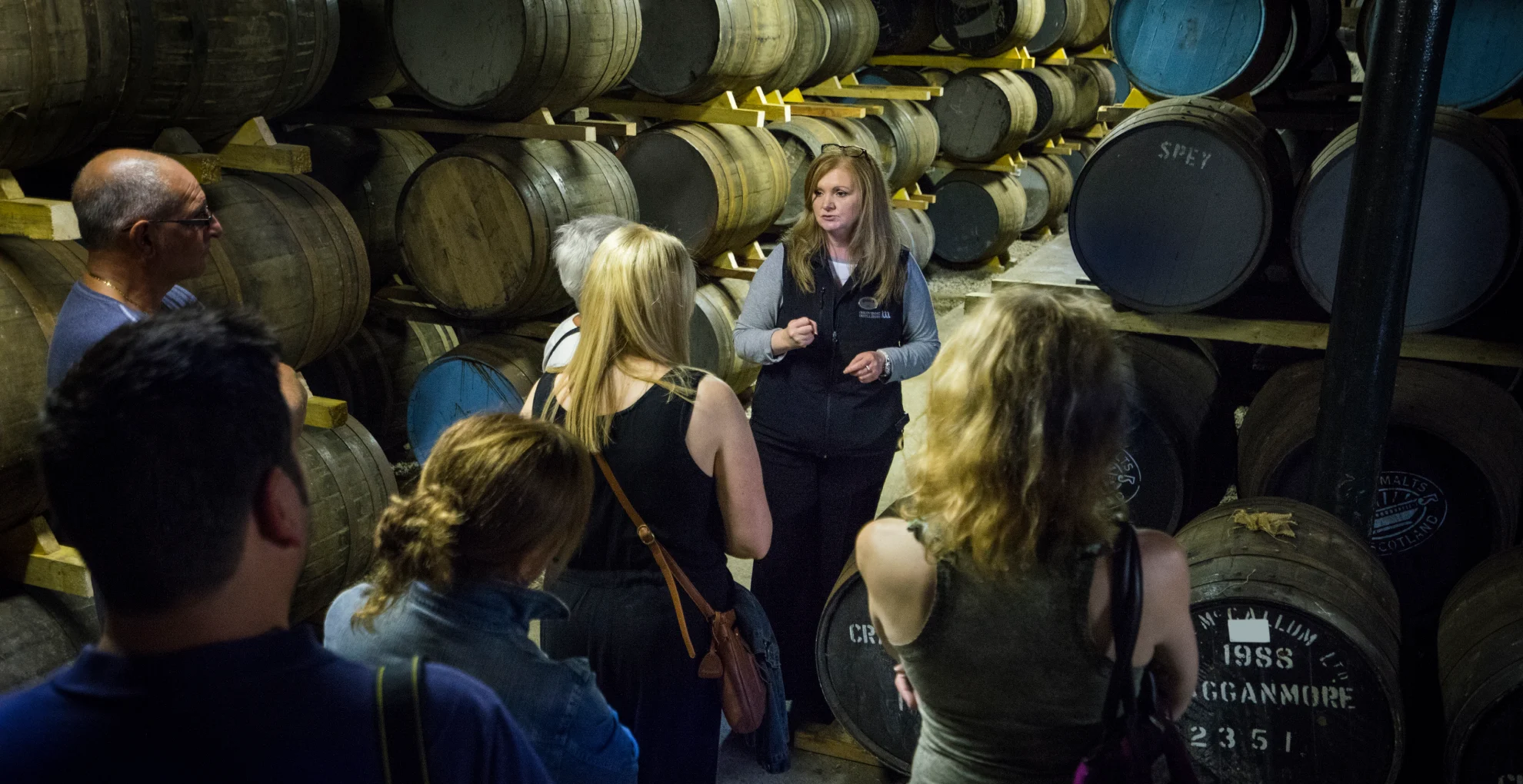 A tour guide stands in a room containing lots of wooden barrels stacked on top of each other. The guide speaks to a group of people who face her.