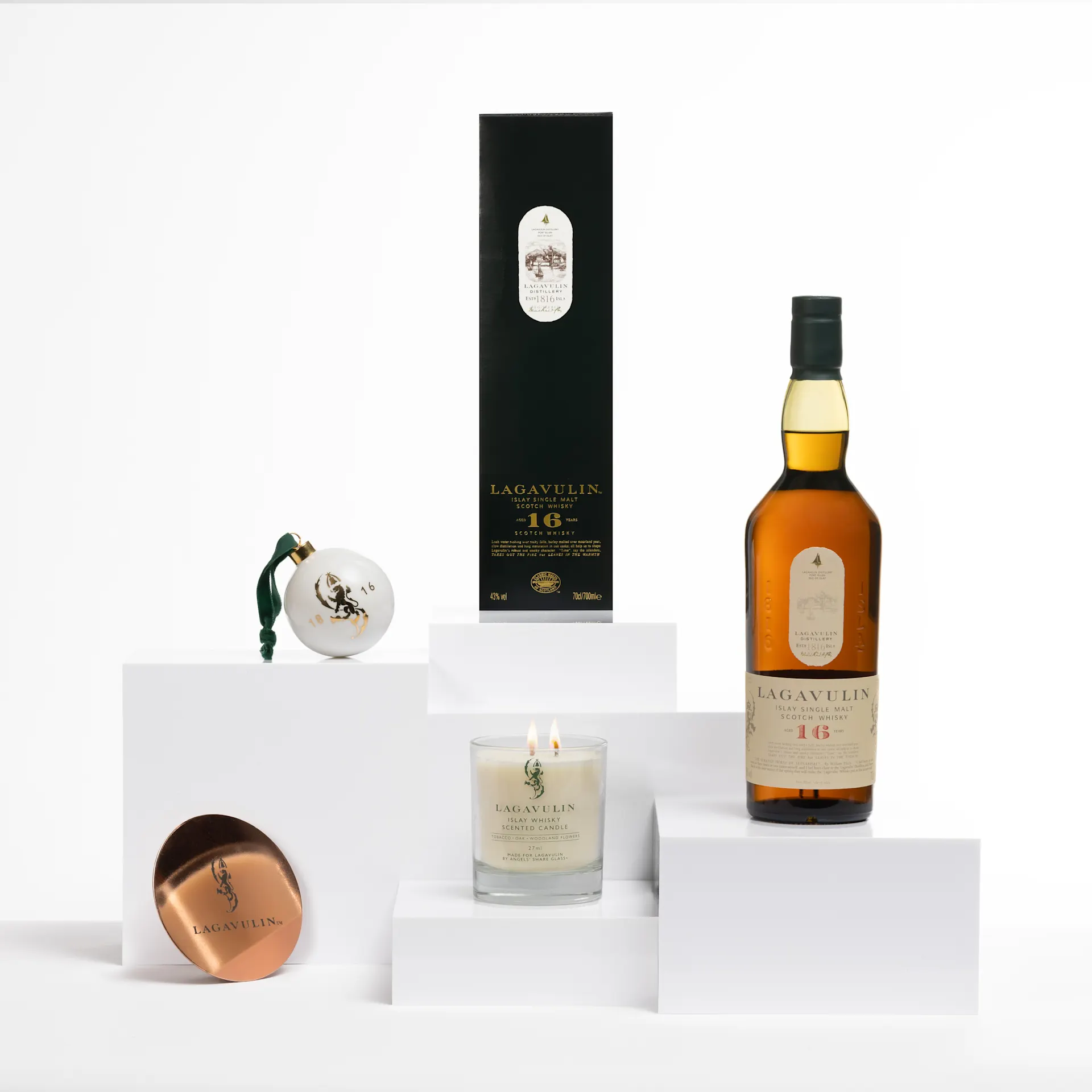 Een Lagavulin 16 fles, een merkbal, een brandende kaars en een koperen onderzetter op witte blokken.