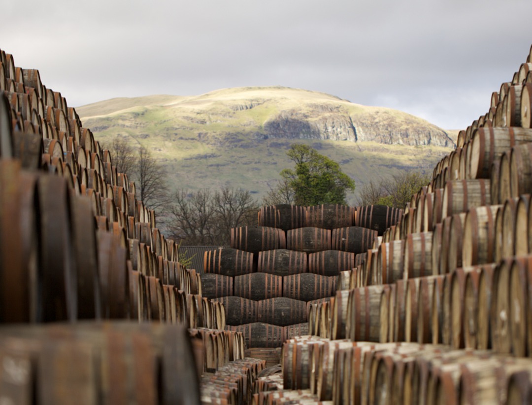 Hundreds of whisky barrels are stacked up, with a large mountain sitting behind them.