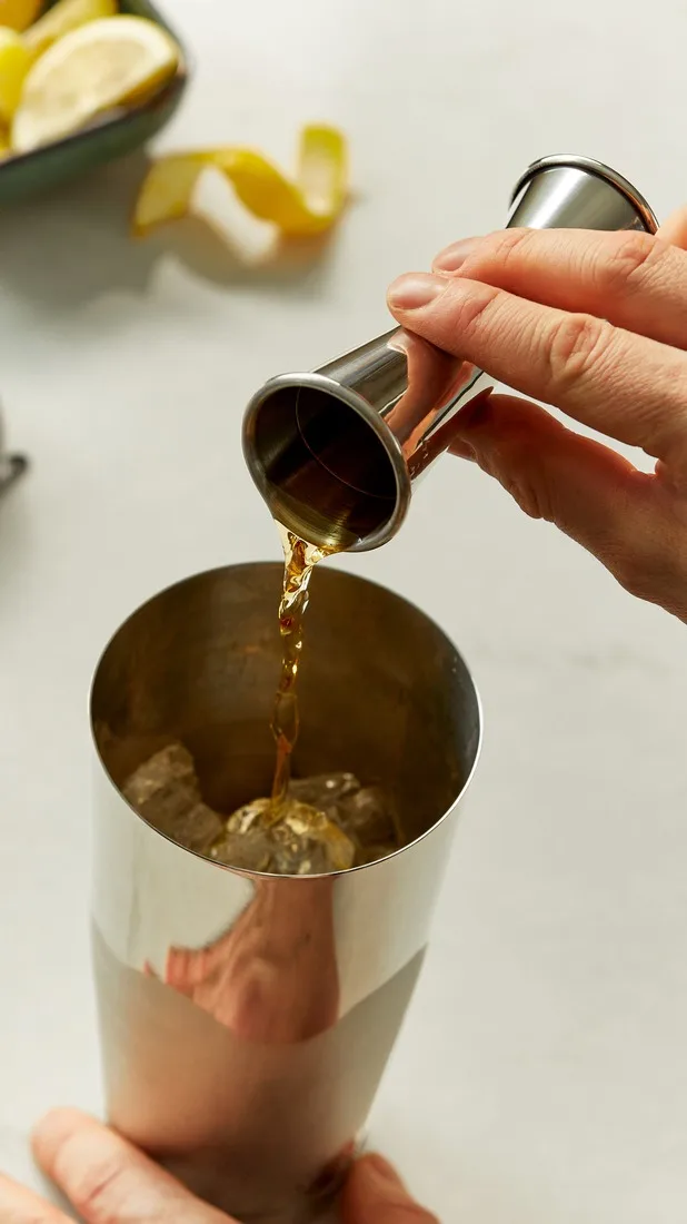 Een jigger giet een donkere sterke drank in een cocktailshaker gevuld met ijs.