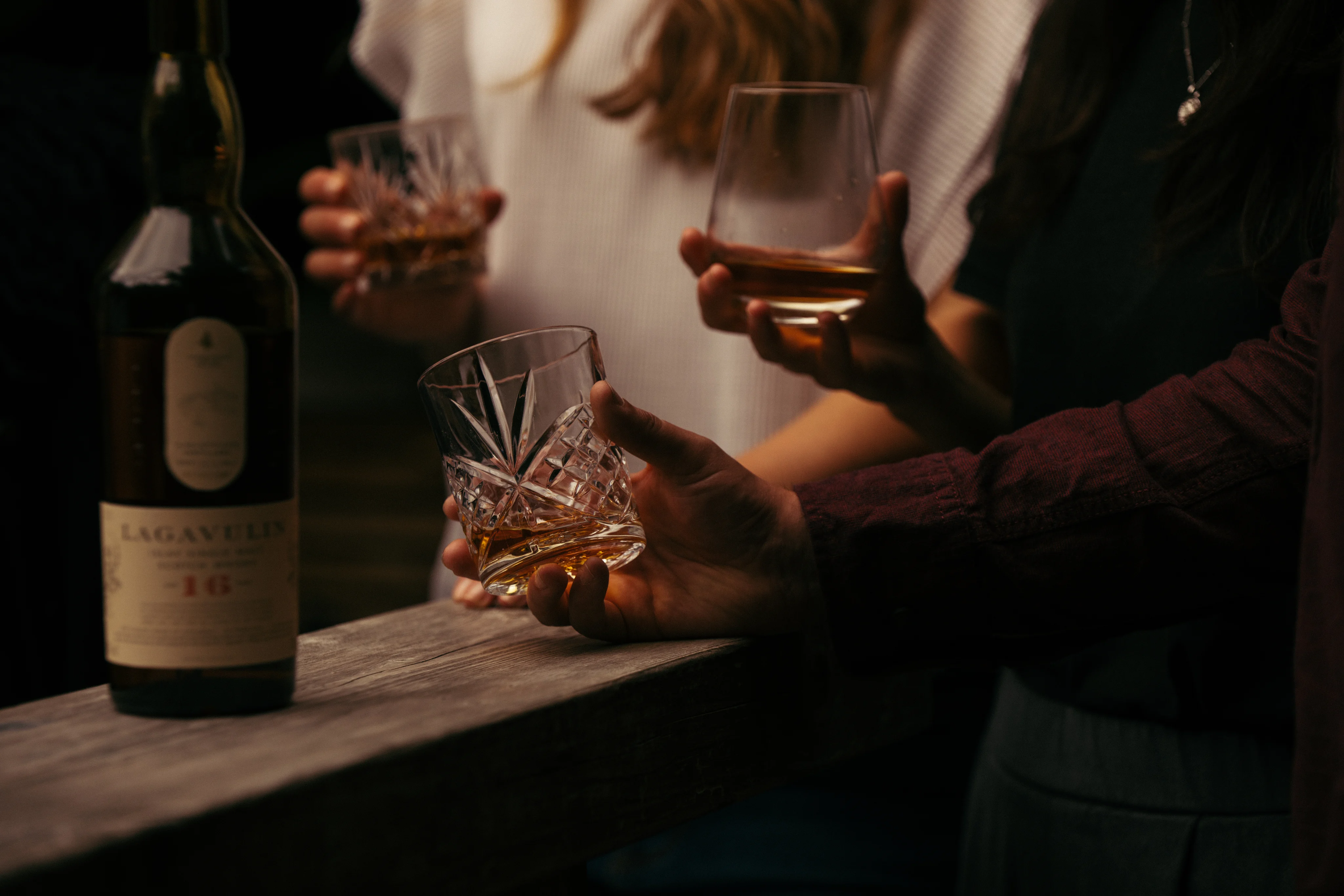 木桌上放著一瓶 Lagavulin 威士卡。酒瓶後面站著一個穿白衣的人，他拿著一個裝有威士忌的石杯。在這個人的前面有兩隻不同的手，各拿著一個裝有威士忌的石杯。 