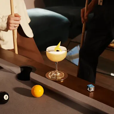 A whisky sour cocktail sits on the edge of a pool table, while a woman stands next to it holding a pool cue.