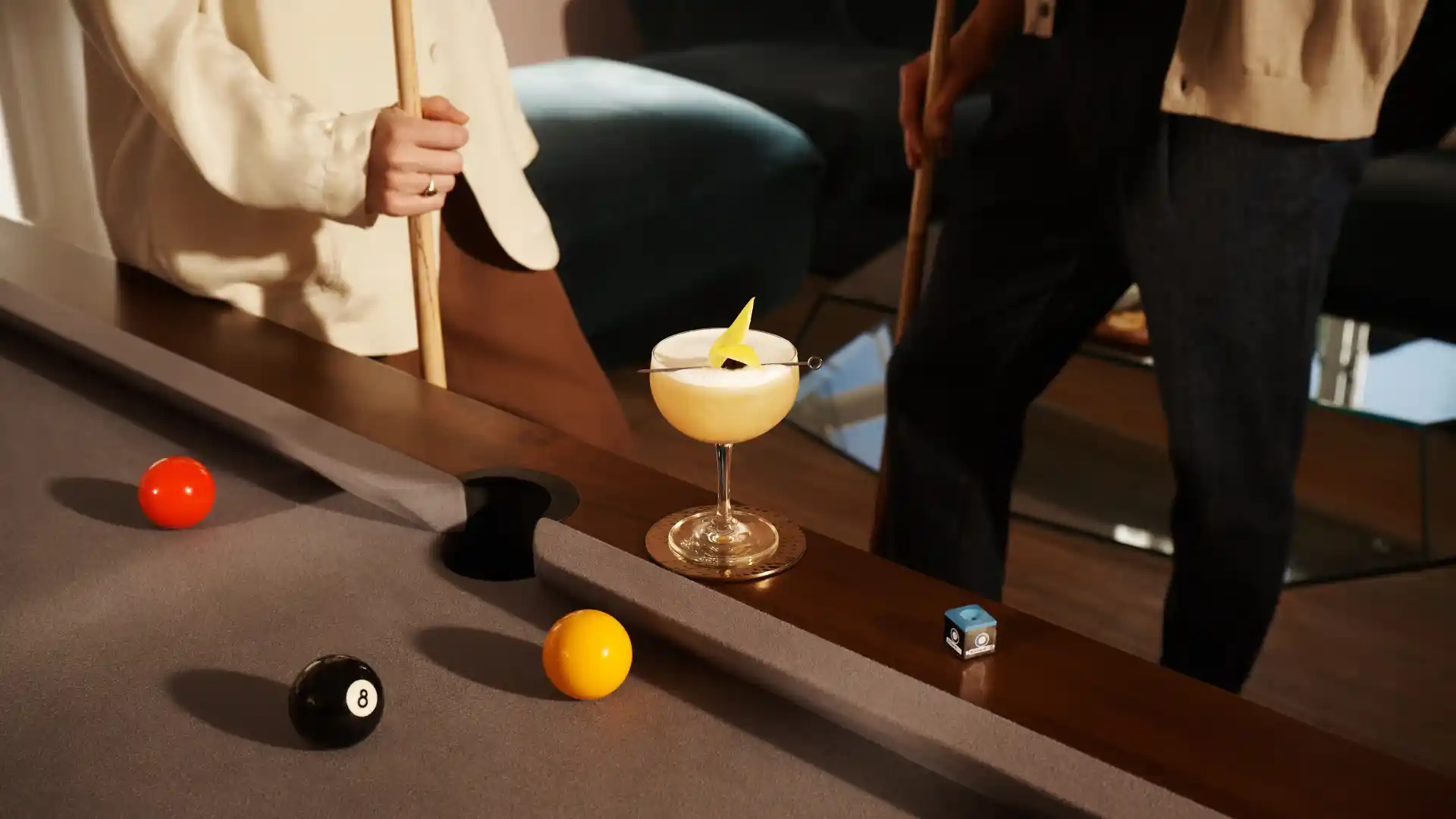 Een whisky sour cocktail staat op de rand van een biljarttafel, terwijl een vrouw ernaast een biljartkeu vasthoudt.