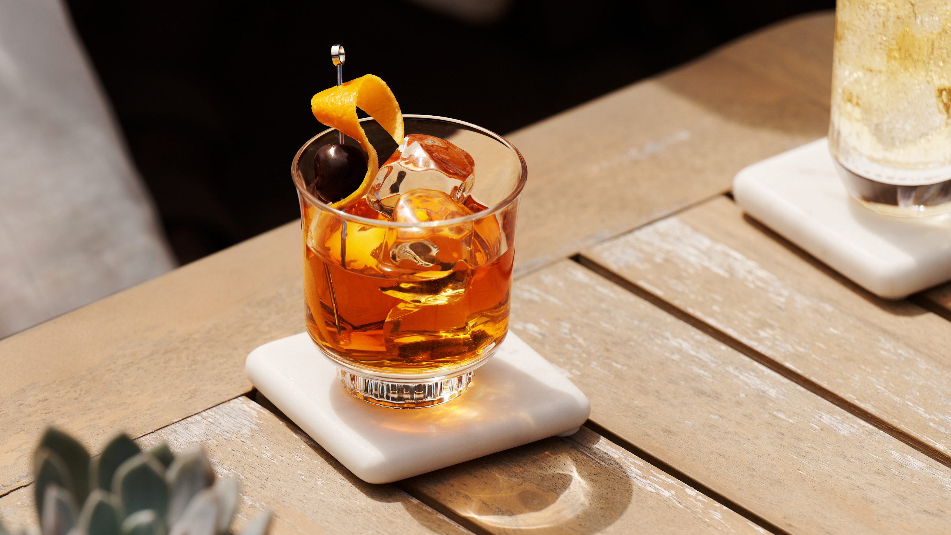 An Old Fashioned cocktail sits in the sun on a wooden surface with a small plant and a bowl of peaches nearby