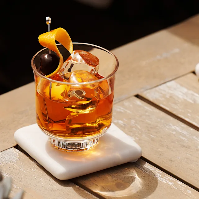 An Old Fashioned cocktail sits in the sun on a wooden surface with a small plant and a bowl of peaches nearby