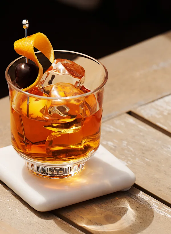 Een Old Fashioned cocktail staat buiten op een houten oppervlak. De garnering bestaat uit een kers en een gedraaide sinaasappelschil.