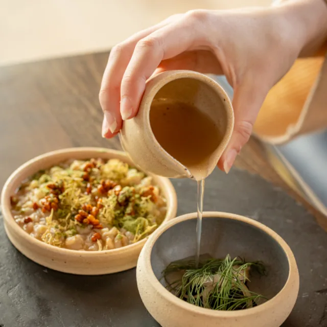 Mehr erfahren Two clay bowl sit on a circular stone disc on a wooden table. The larger bowl contains risotto, and the smaller bowl contains onions and herbs. A hand holds a small clay jug above the smaller bowl and pours a liquid into it.