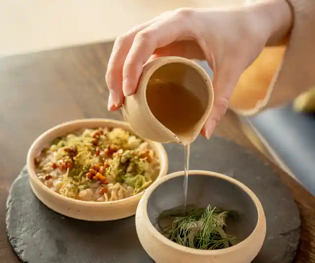 Two clay bowl sit on a circular stone disc on a wooden table. The larger bowl contains risotto, and the smaller bowl contains onions and herbs. A hand holds a small clay jug above the smaller bowl and pours a liquid into it.