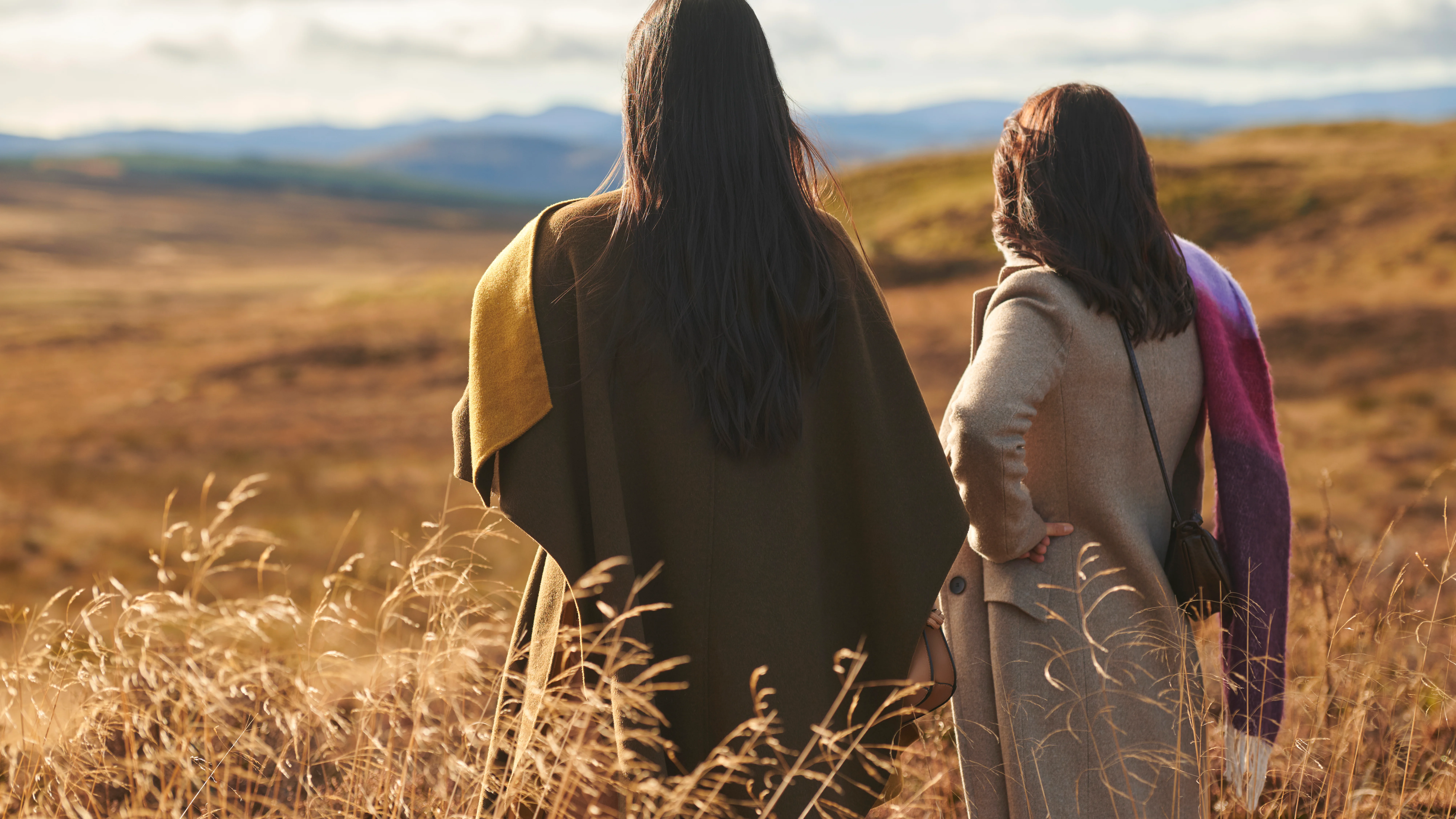 Two friends look out over the Highlands in autumn. In the background are several hills covered in golden grass.