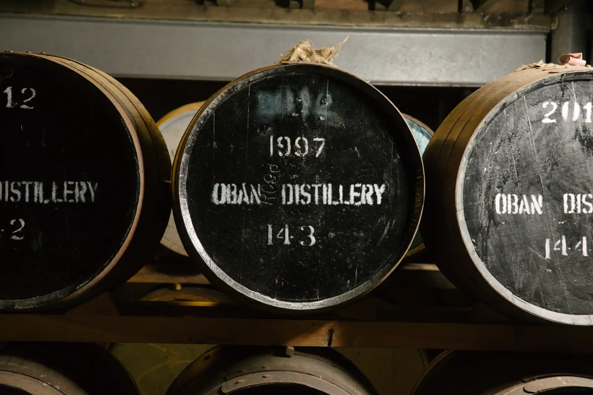 Wooden barrels sit in a row of 3 by 2. The front of each barrel is painted black with white writing printed on stating ‘1997 Oban Distillery’.