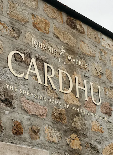 Auf einer Backsteinmauer ist ein goldenes Schild mit der Aufschrift Cardhu zu sehen, und an der Mauer steht eine schwarze Laterne.