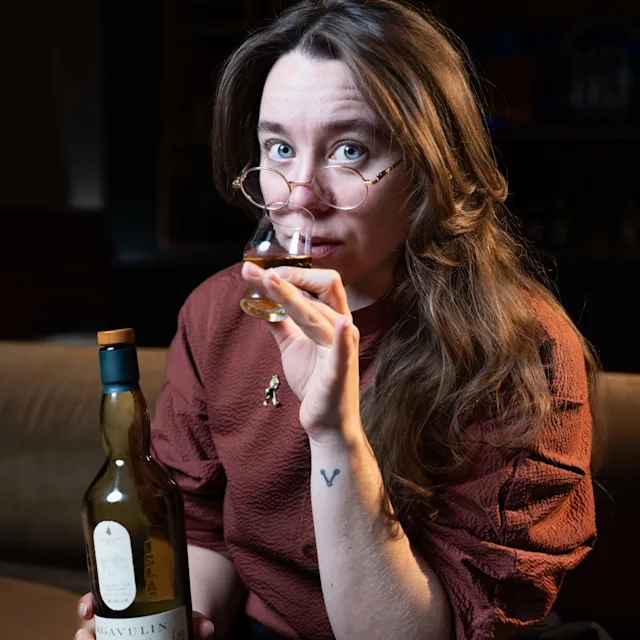 Charlotte Barker holds a whisky tasting glass and holds it to her lips. In her other hand she holds a bottle of Lagavulin 16 year old single malt whisky.