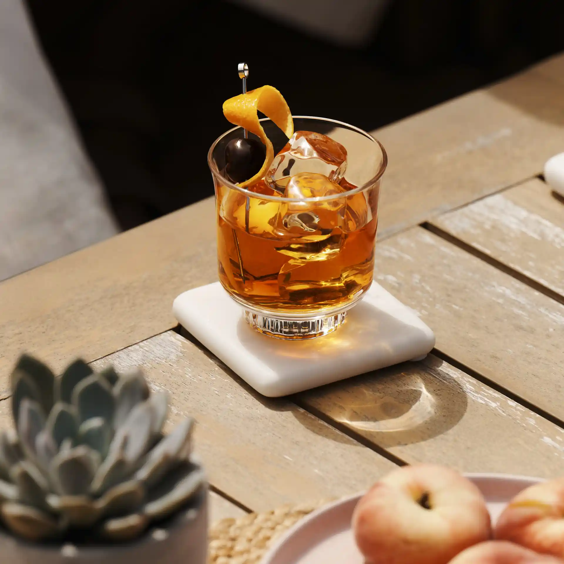 An Old Fashioned cocktail sits on a wooden surface, with a small plant and bowl of peaches nearby.