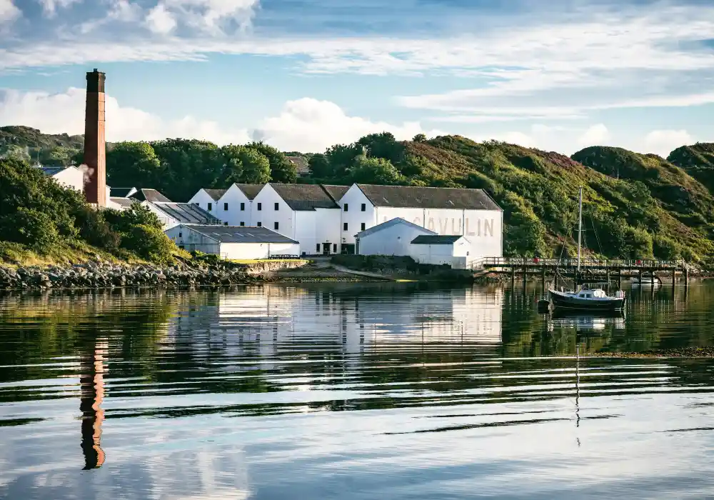 The while buildings of Lagavulin distillery sit next to a calm body of water, on which stands a pier and a small boat. Behind the distillery are large green hills.  