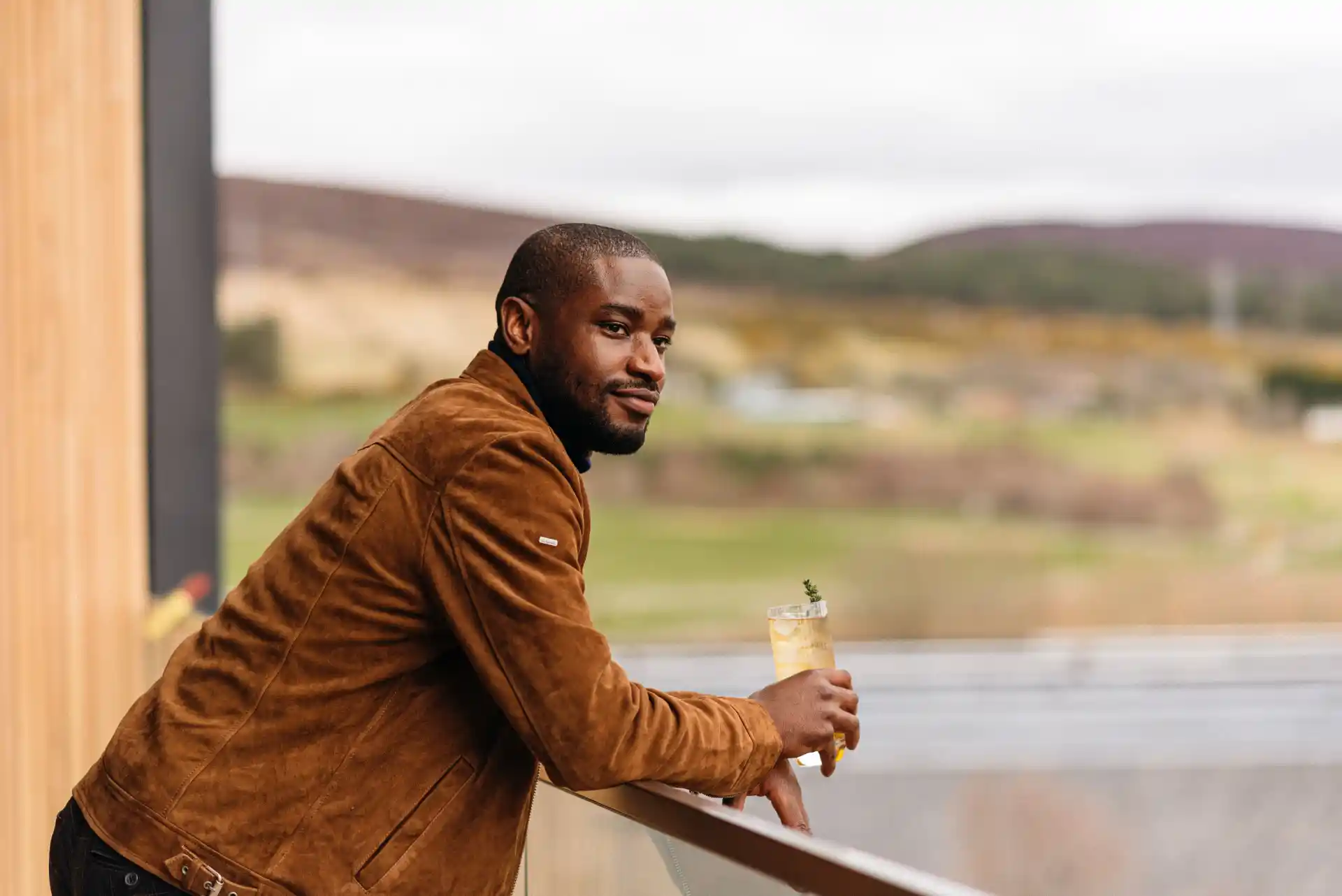 Een man leunt over een balkon en kijkt uit over het landschap. Hij houdt een glas met een whiskycocktail, ijs en een takje rozemarijn vast.