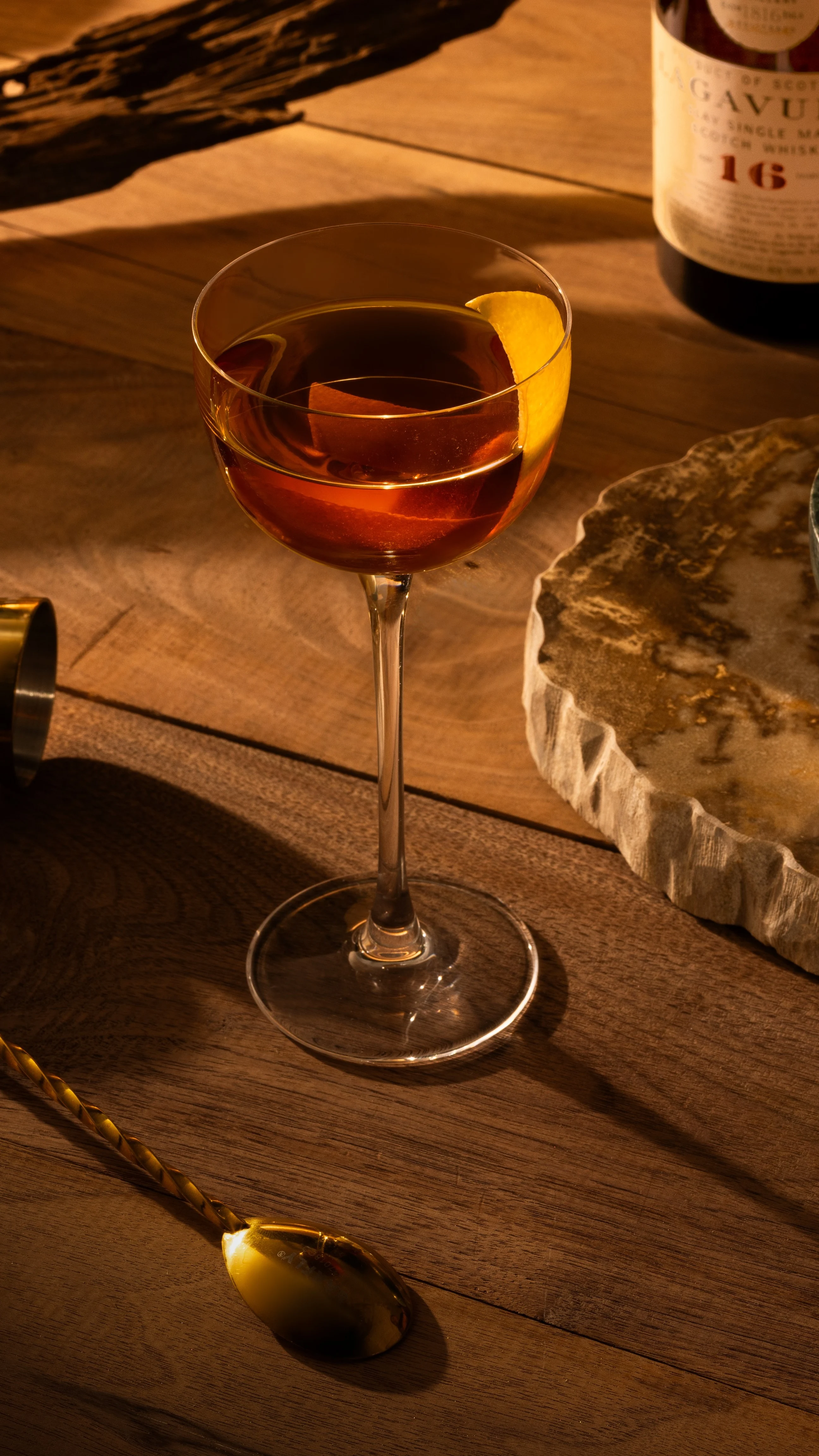 A Peaty Burns cocktail in a coupe glass on a wooden surface surrounded by cocktail-making equipment.