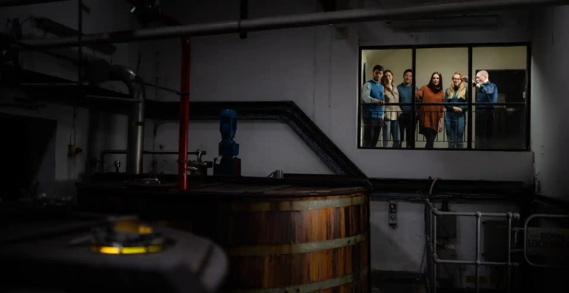 A tour guide and tour group stand on a balcony, overlooking a large whisky mash tun.