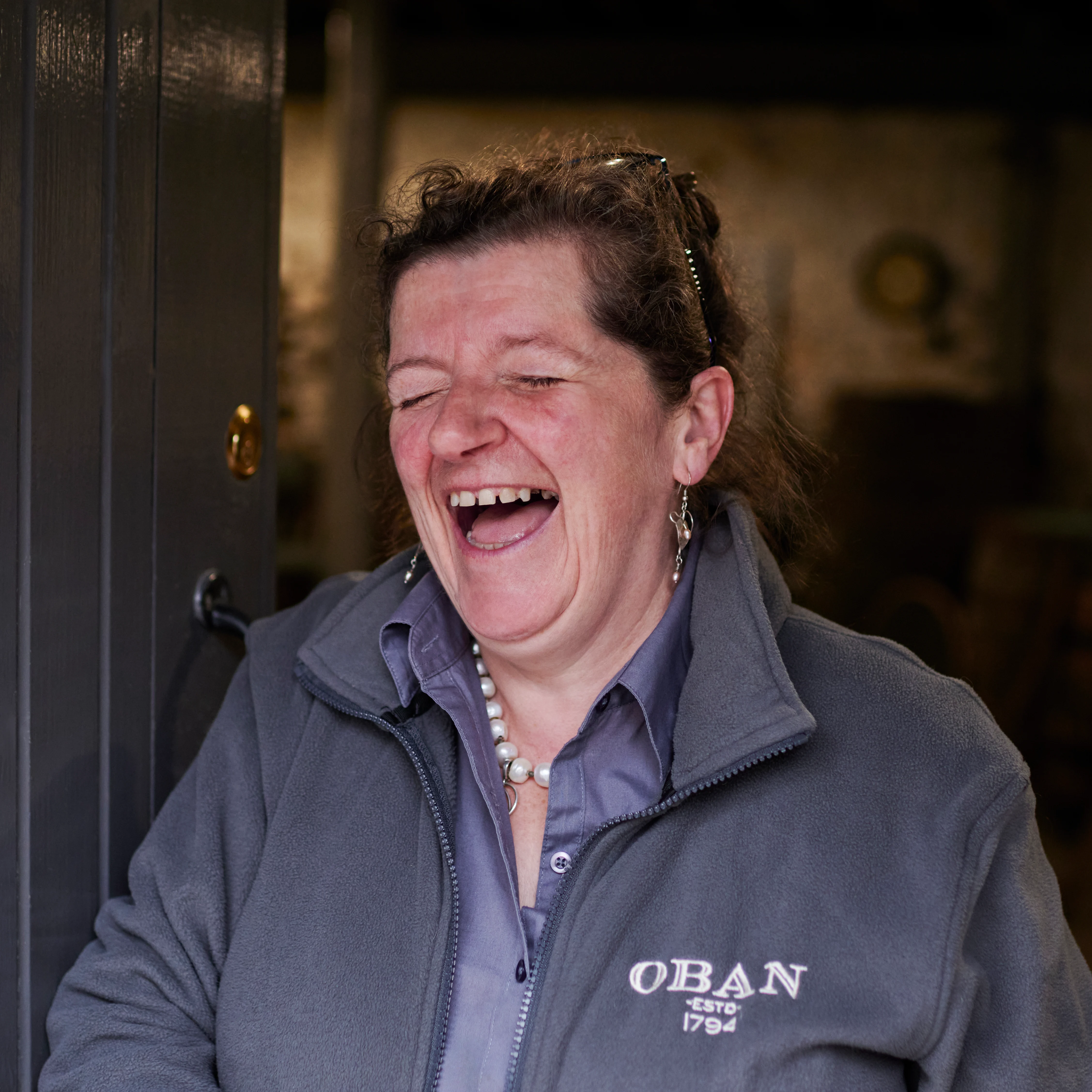 A woman laughing and smiling during a guided whisky distillery tour at Oban, wearing a navy jacket with distillery signage visible behind her