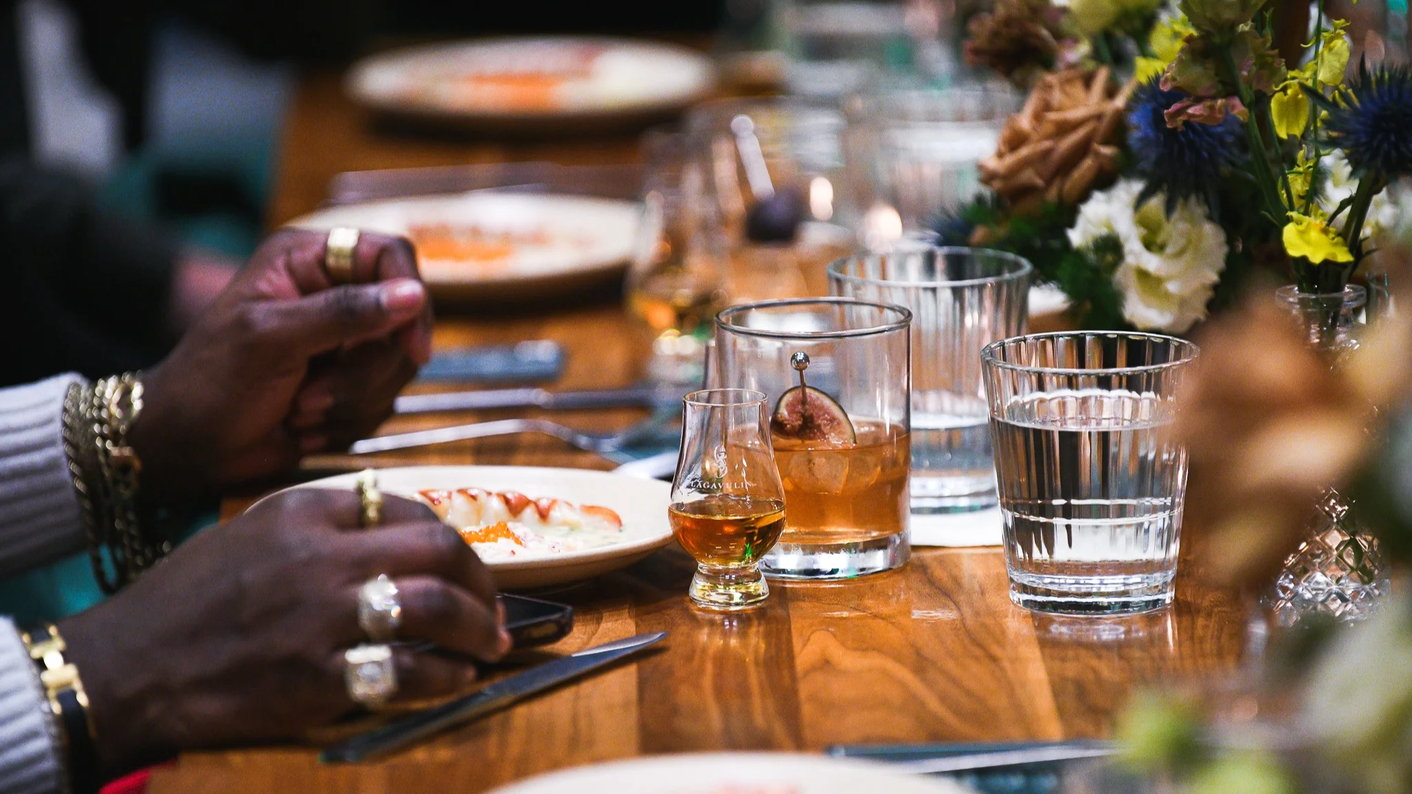 The hands of a man with gold jewellery at a lively dinner party, with whisky glasses on the table. 