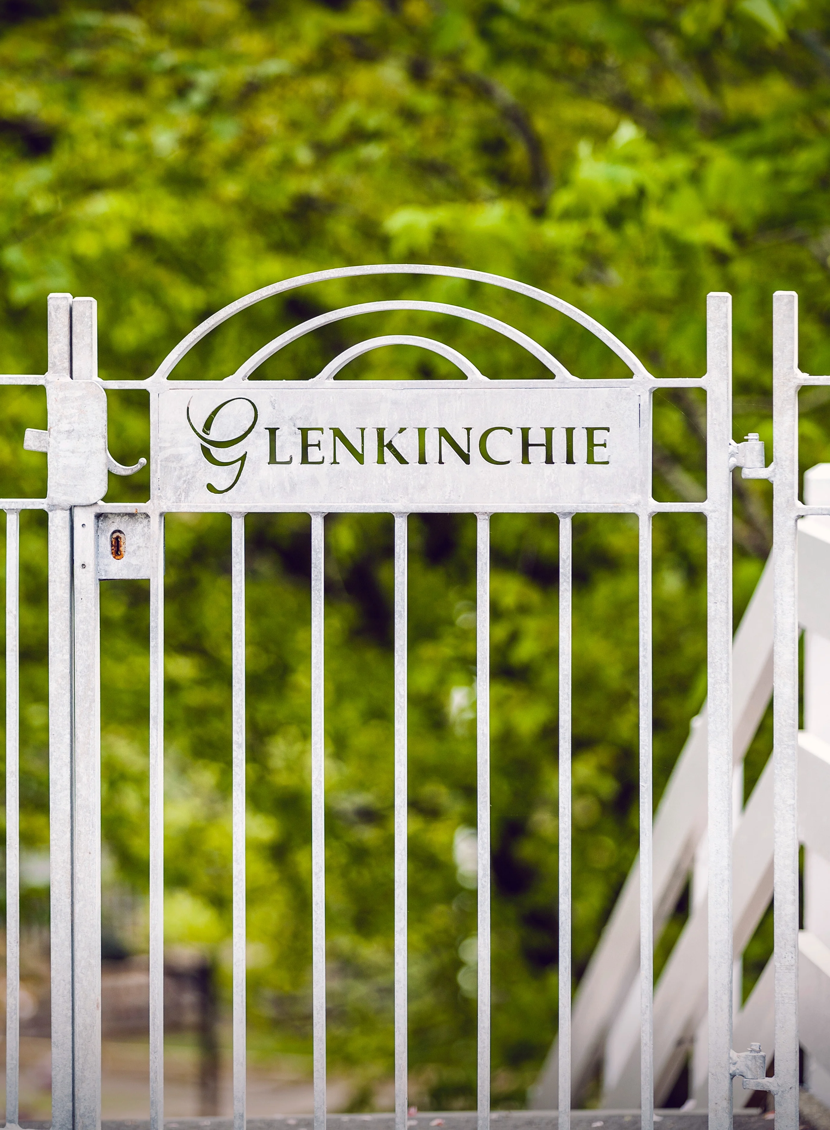 Een wit metalen hek met het opschrift „Glenkinchie” tegen een achtergrond van groene bomen.