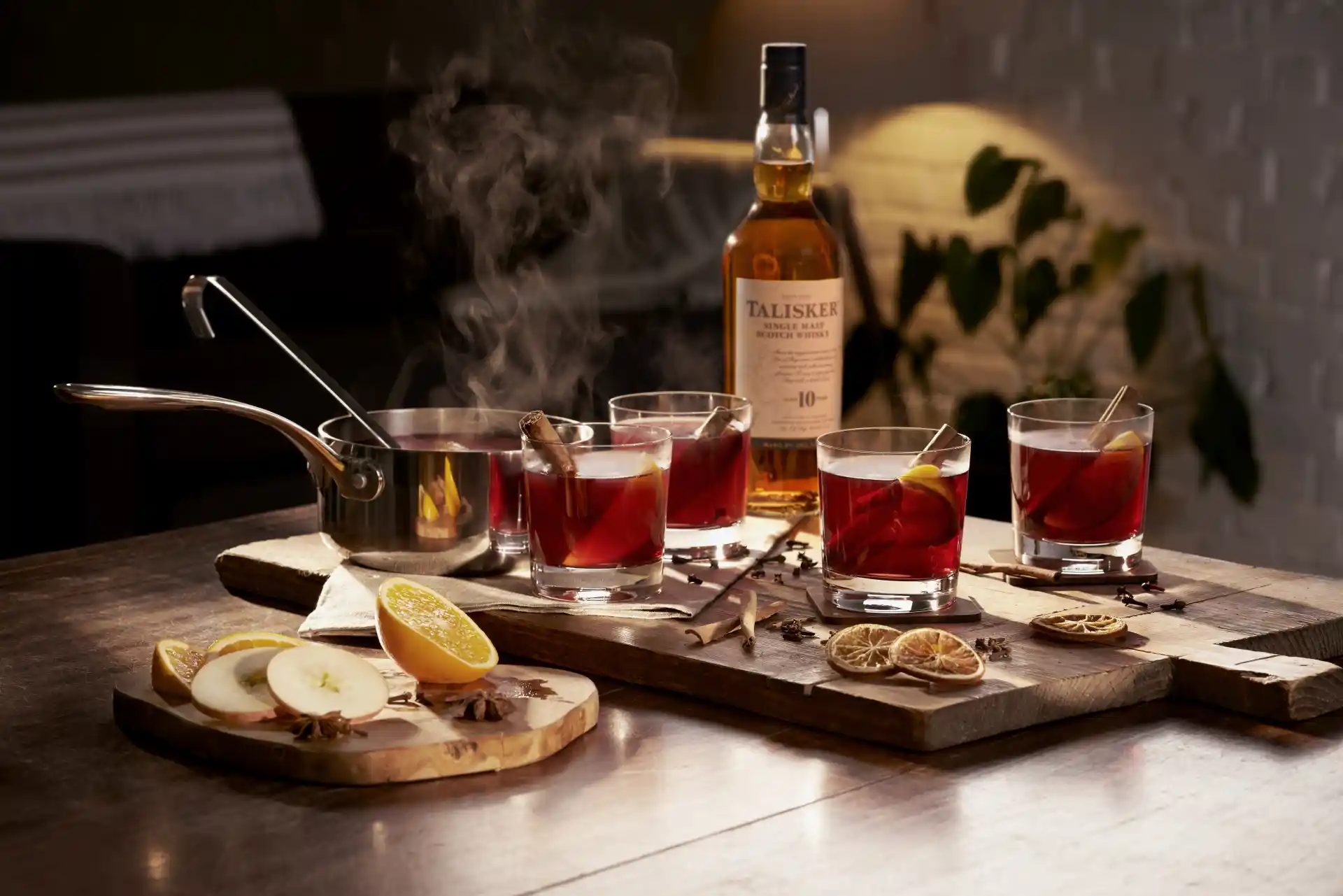 A bottle of Talisker 10 sits on a wooden board, surrounded by a saucepan and four rocks glasses containing hot toddies.