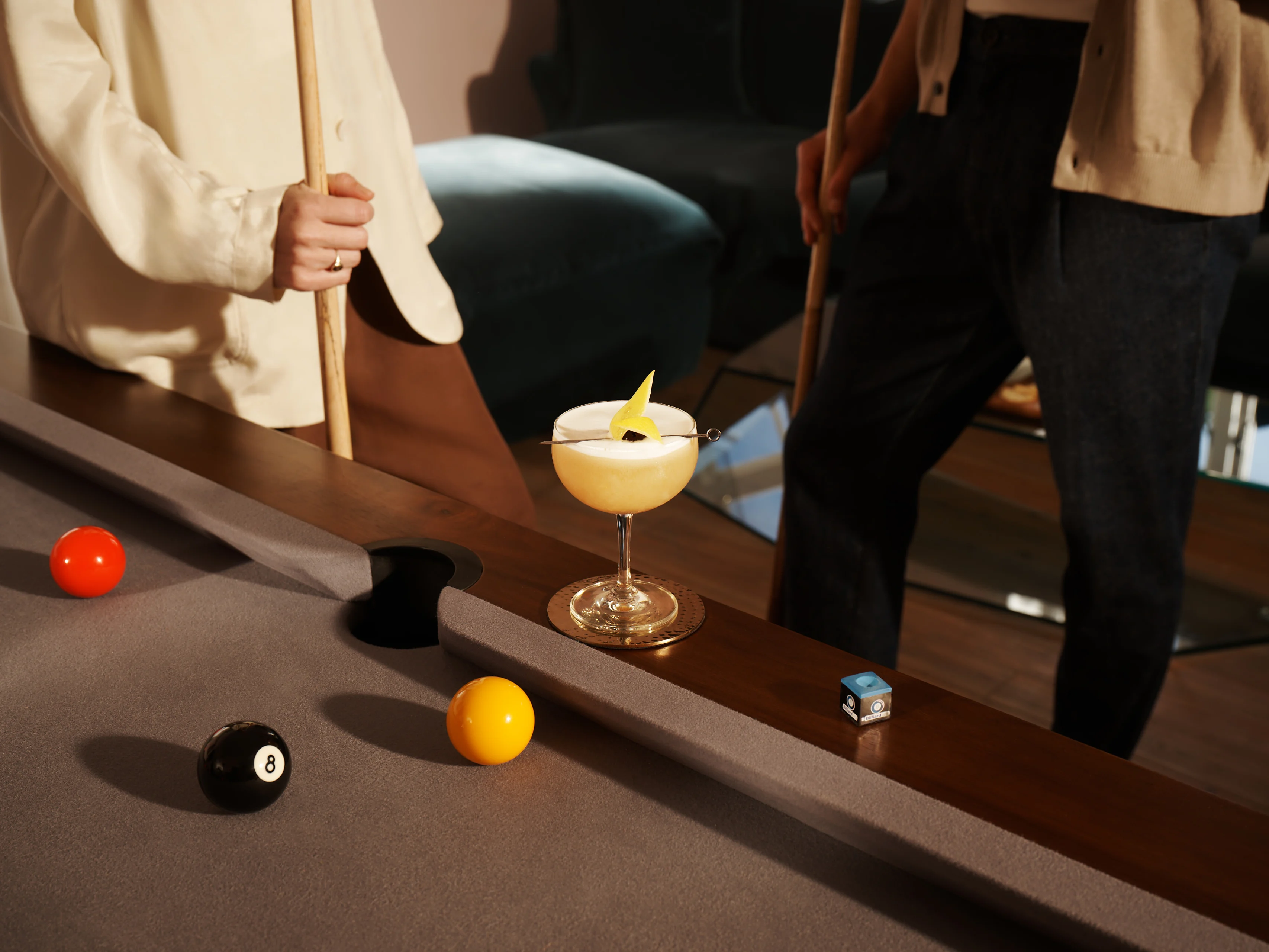 Ein Whisky-Sour-Cocktail liegt auf der Kante eines Billardtisches, während eine Frau daneben steht und einen Billardqueue hält.