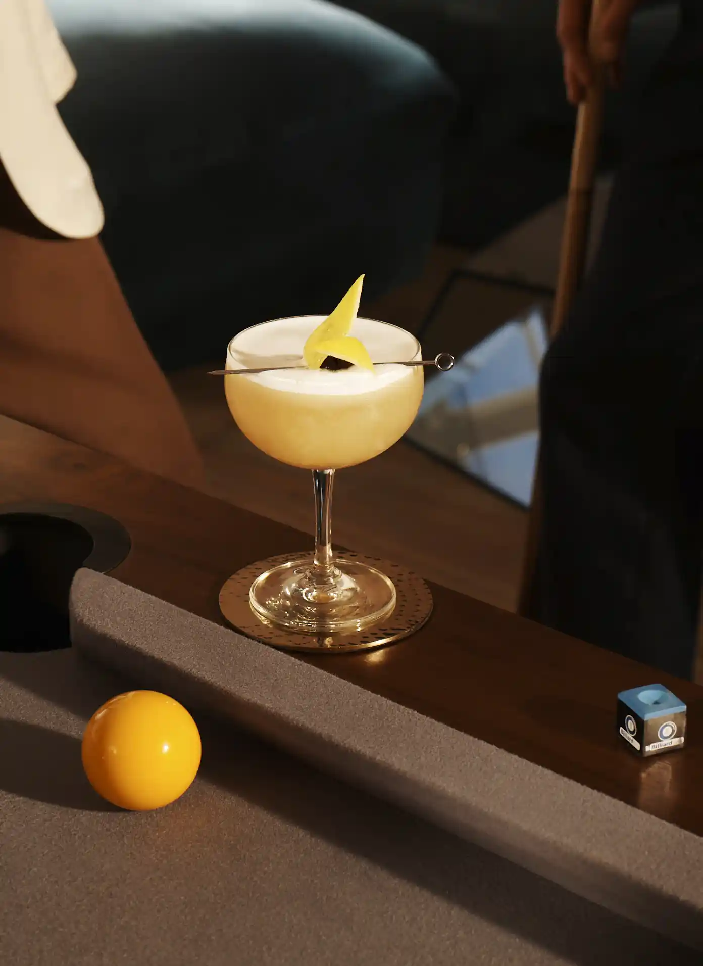 A whisky sour cocktail sits on the edge of a pool table, while a woman stands next to it holding a pool cue.