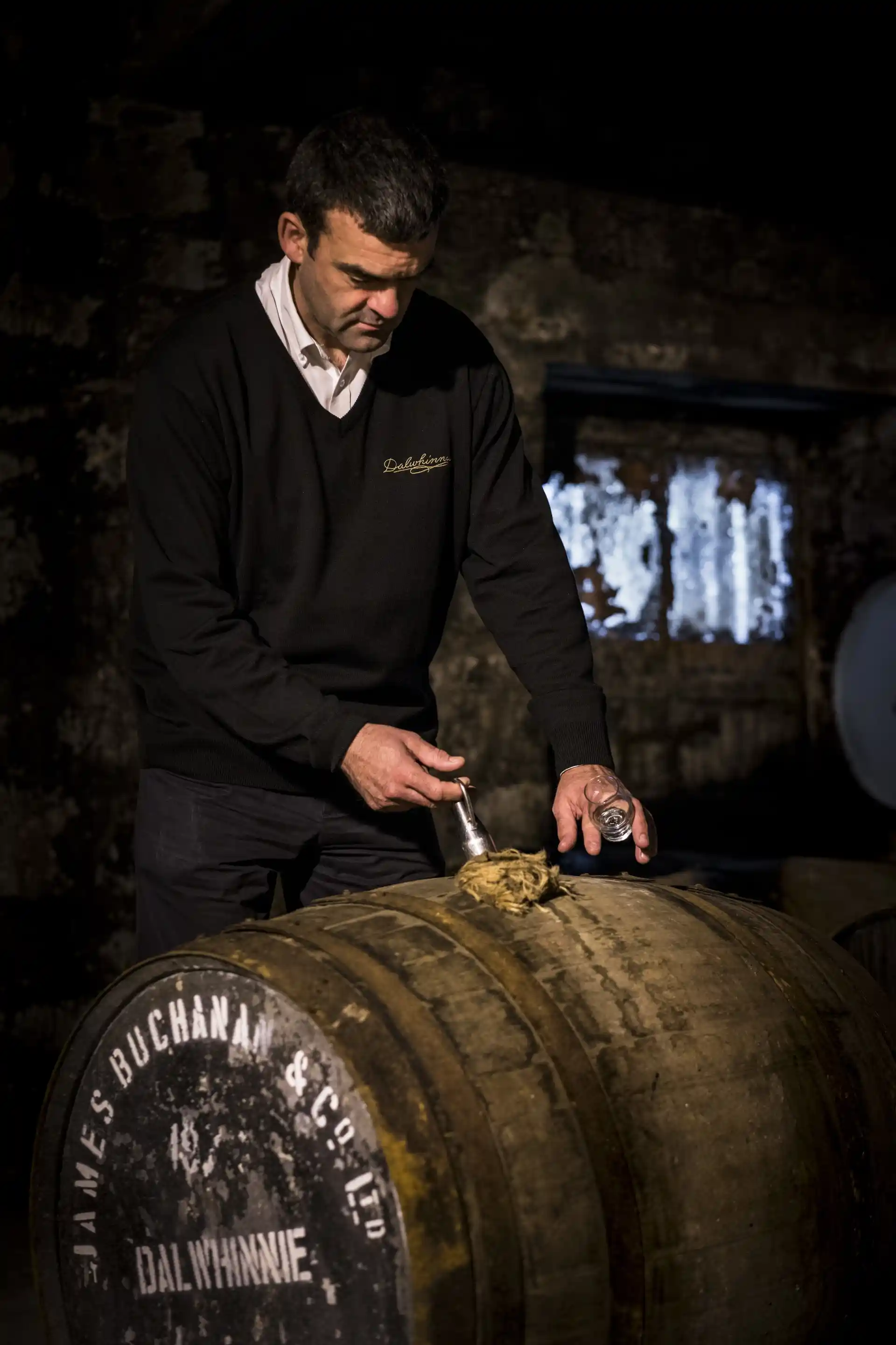 Een medewerker van Dalwhinnie gebruikt een metalen pipet om whisky uit een houten vat te halen dat op zijn kant ligt. In de andere hand van de medewerker staat een leeg dramglas.