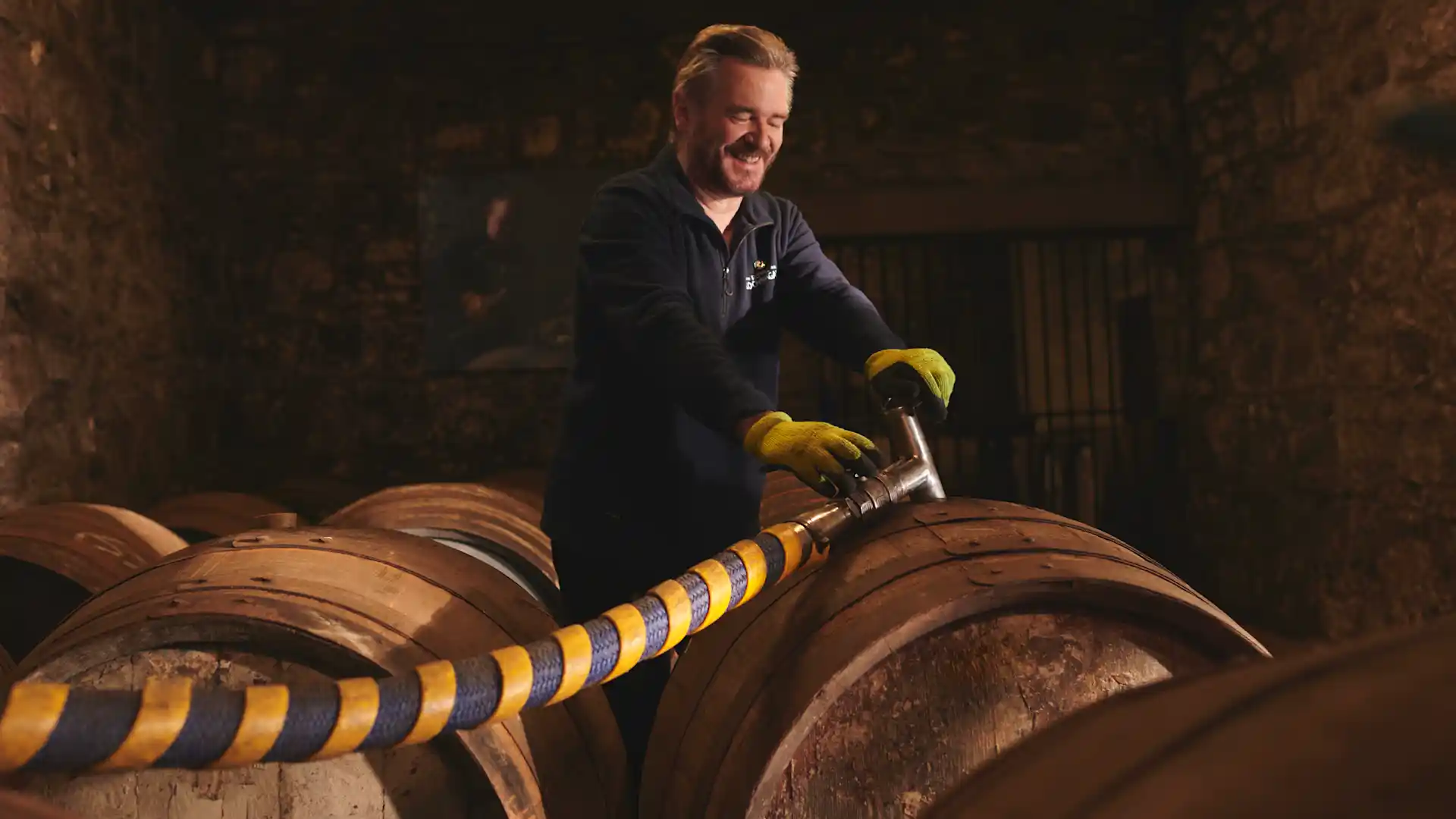 A distillery worker stands in a room of whisky barrels. He fills one barrel with whisky, using a long hose with a silver nozzle.