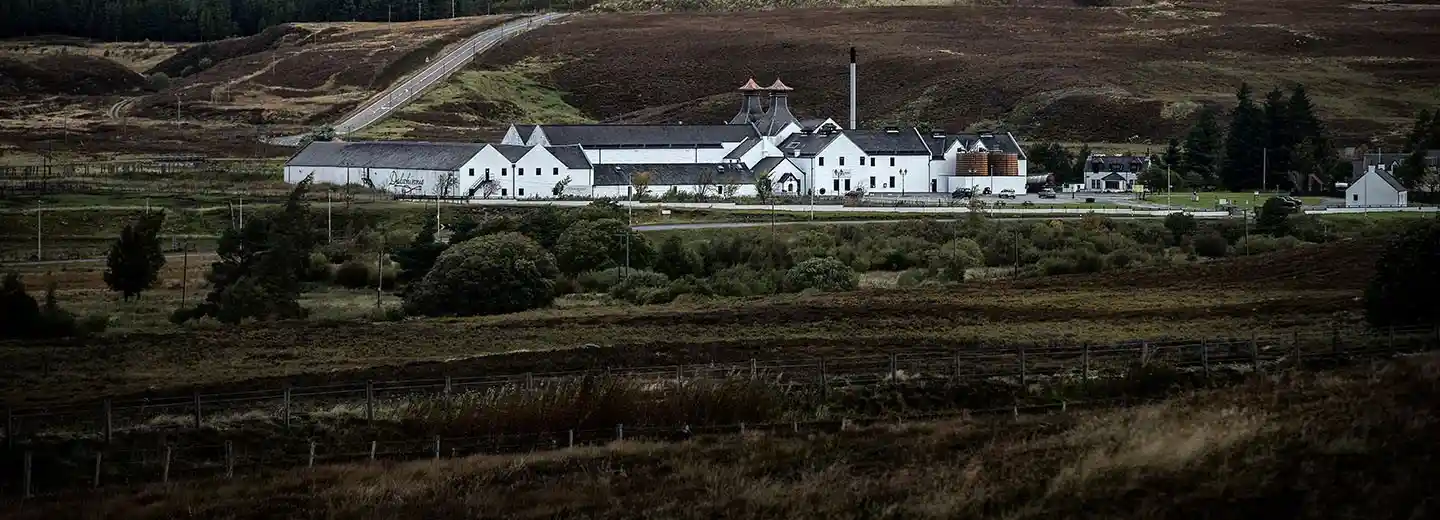 The white buildings of Dalwhinnie distillery are seen from a distance. In front of the buildings are dark green fields and trees. Behind the distillery are brown and green hills.