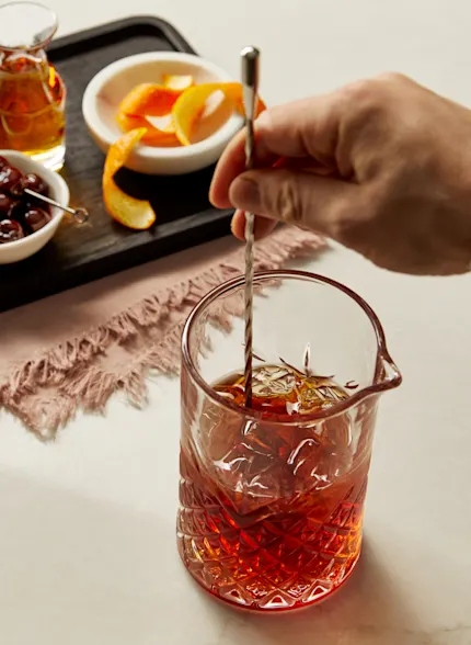 Eine Person rührt eine bernsteinfarbene Flüssigkeit in einem geschliffenen Kristallglas auf einer Küchenfläche um. Auf der Oberfläche stehen auch eine Schale mit Orangenschalen und Maraschino-Kirschen.