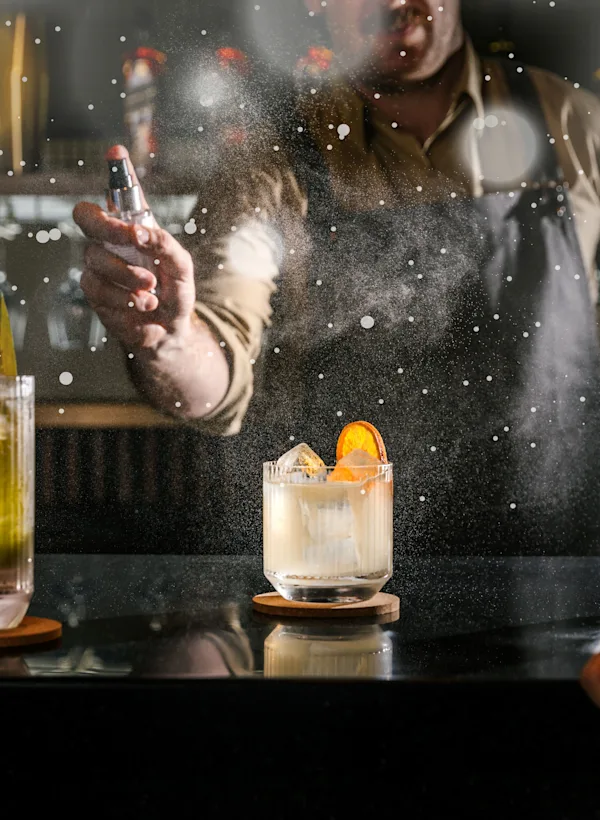Two cocktails and a glass of water placed in a bar counter, with a barman in the background