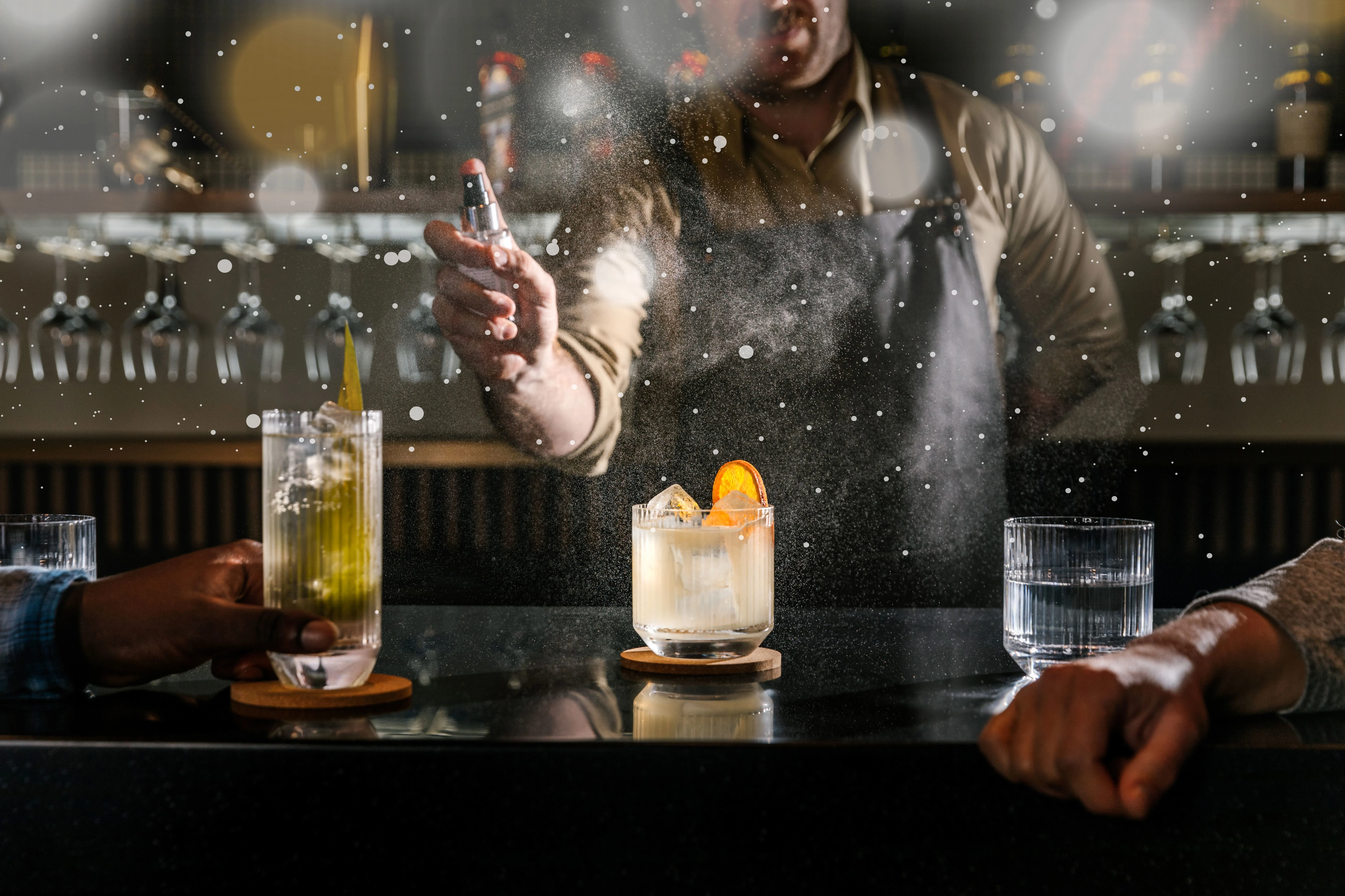 Two cocktails and a glass of water placed in a bar counter, with a barman in the background 