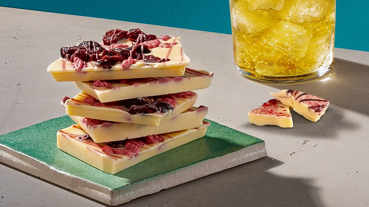 Ein gelber Whisky-Cocktail neben einem Teller mit einem Turm aus weißer Schokolade mit Himbeeren.