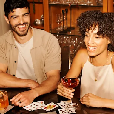 A man and woman sit at a table with playing cards and whisky cocktails