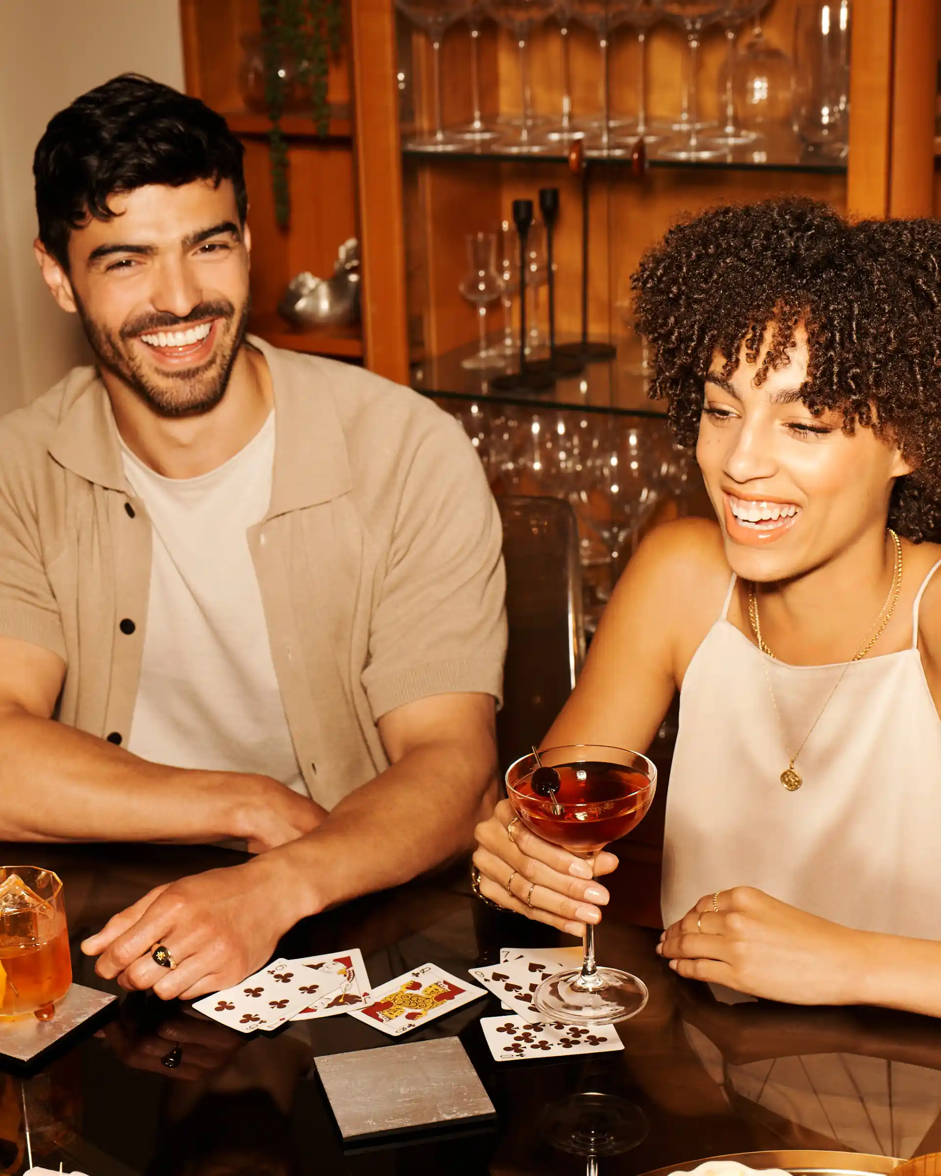 Een man en twee vrouwen zitten aan een tafel. Voor hen staan whiskycocktails en speelkaarten.