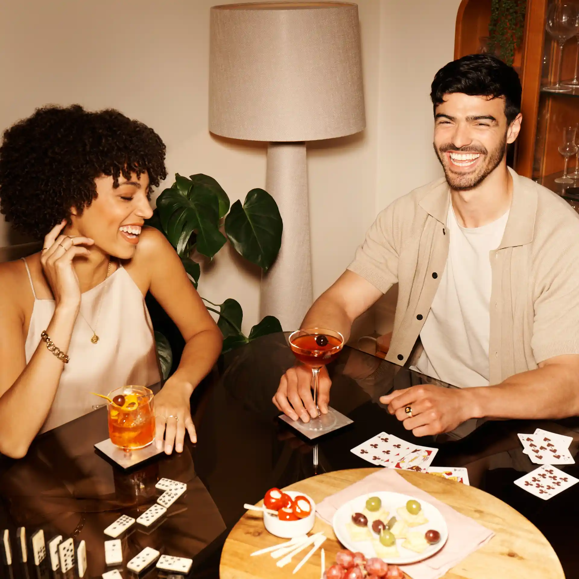 Een man en een vrouw zitten aan een tafel met verschillende whiskycocktails, snacks, dominostenen en speelkaarten.