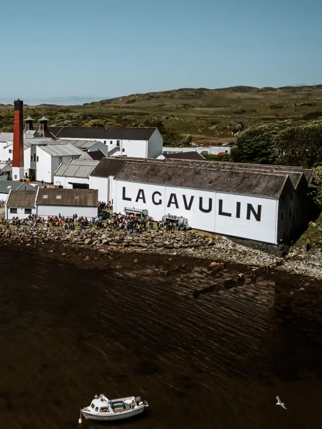 Eine Luftaufnahme der Lagavulin Destillerie auf Islay, Schottland. Das weiße Gebäude mit dem großen "LAGAVULIN" Schriftzug steht direkt an der Küste. Eine Menschenmenge versammelt sich vor der Destillerie, während Boote am Pier anlegen.