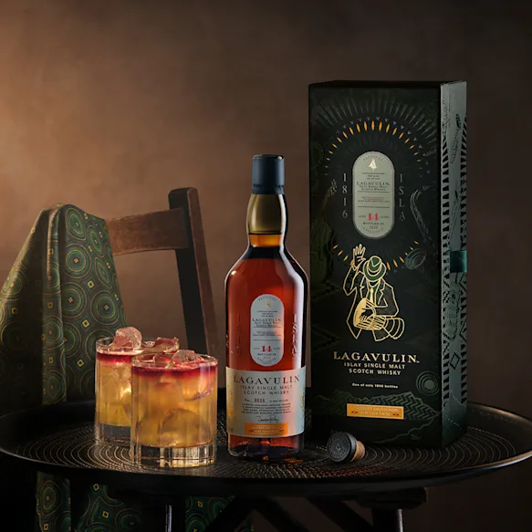 Die Flasche Lagavulin 14 Jahre Islay Single Malt Scotch Whisky und ihre Verpackung. Daneben werden auf einem dunklen Tisch zwei Whisky-Cocktails mit Eis serviert.