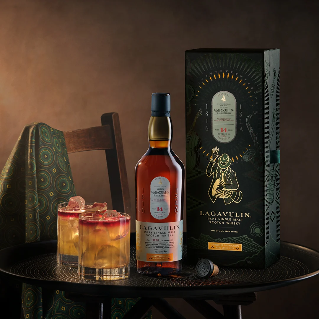 Die Flasche Lagavulin 14 Jahre Islay Single Malt Scotch Whisky und ihre Verpackung. Daneben werden auf einem dunklen Tisch zwei Whisky-Cocktails mit Eis serviert.