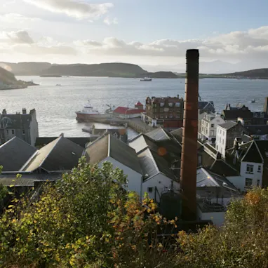 A view of Oban distillery from above, showing the foliage and buildings surrounding the distillery. The distillery overlooks a calm body of water with hills beyond the water. The sky is blue with white clouds.
