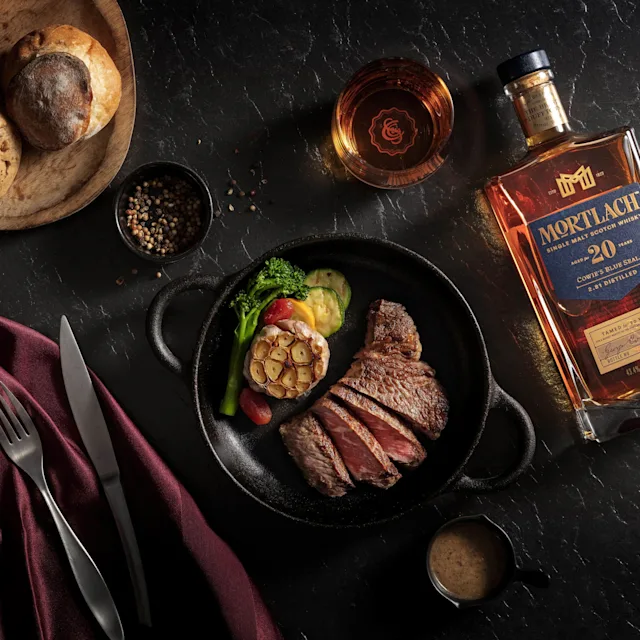 一瓶莫特拉赫16年威士忌、一杯威士忌和一道擺盤精美的菜餚,配有醬汁和綠色蔬菜,整齊地擺放在桌子上,旁邊有一束花和餐具。