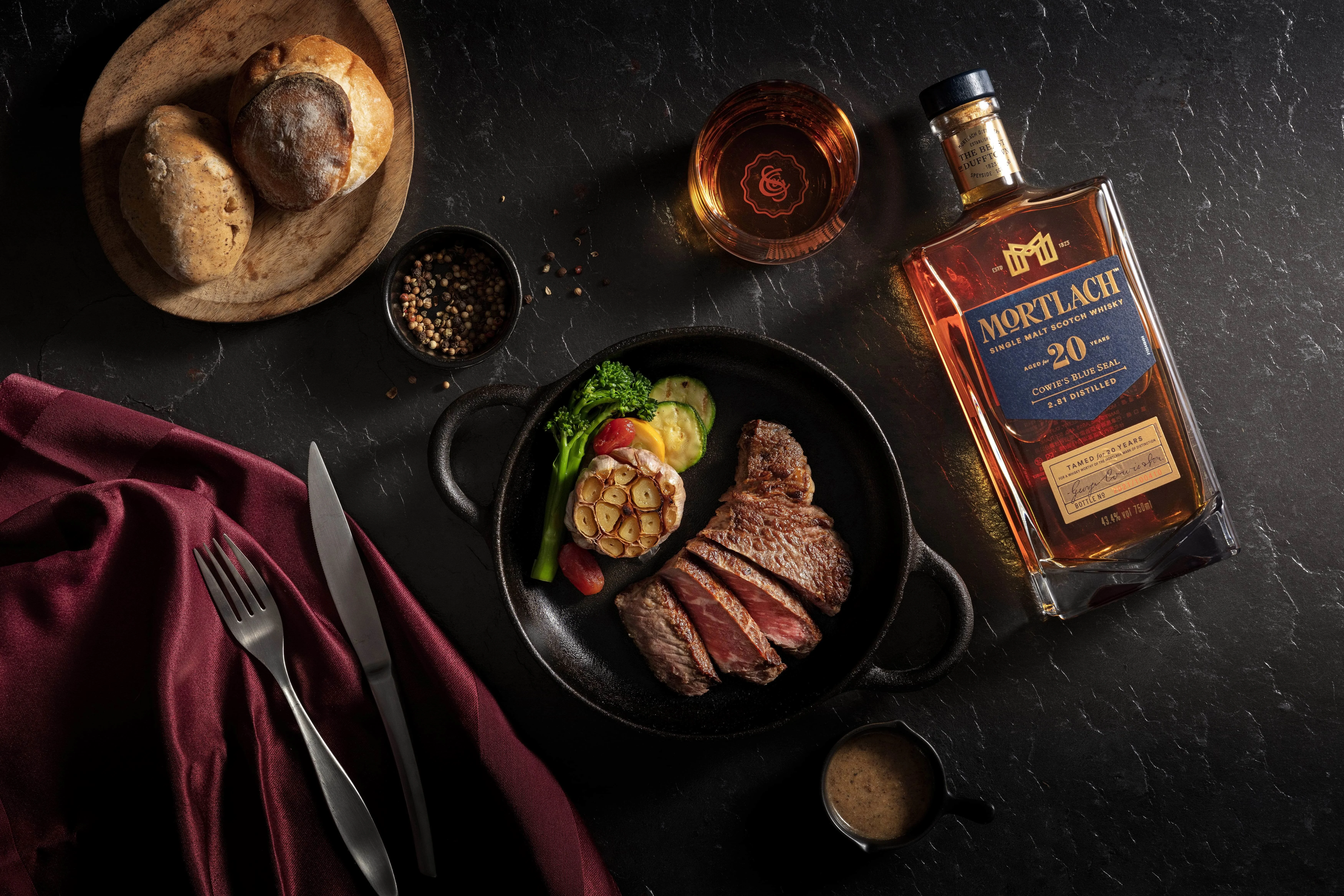 一瓶莫特拉赫16年威士忌、一杯威士忌和一道擺盤精美的菜餚,配有醬汁和綠色蔬菜,整齊地擺放在桌子上,旁邊有一束花和餐具。