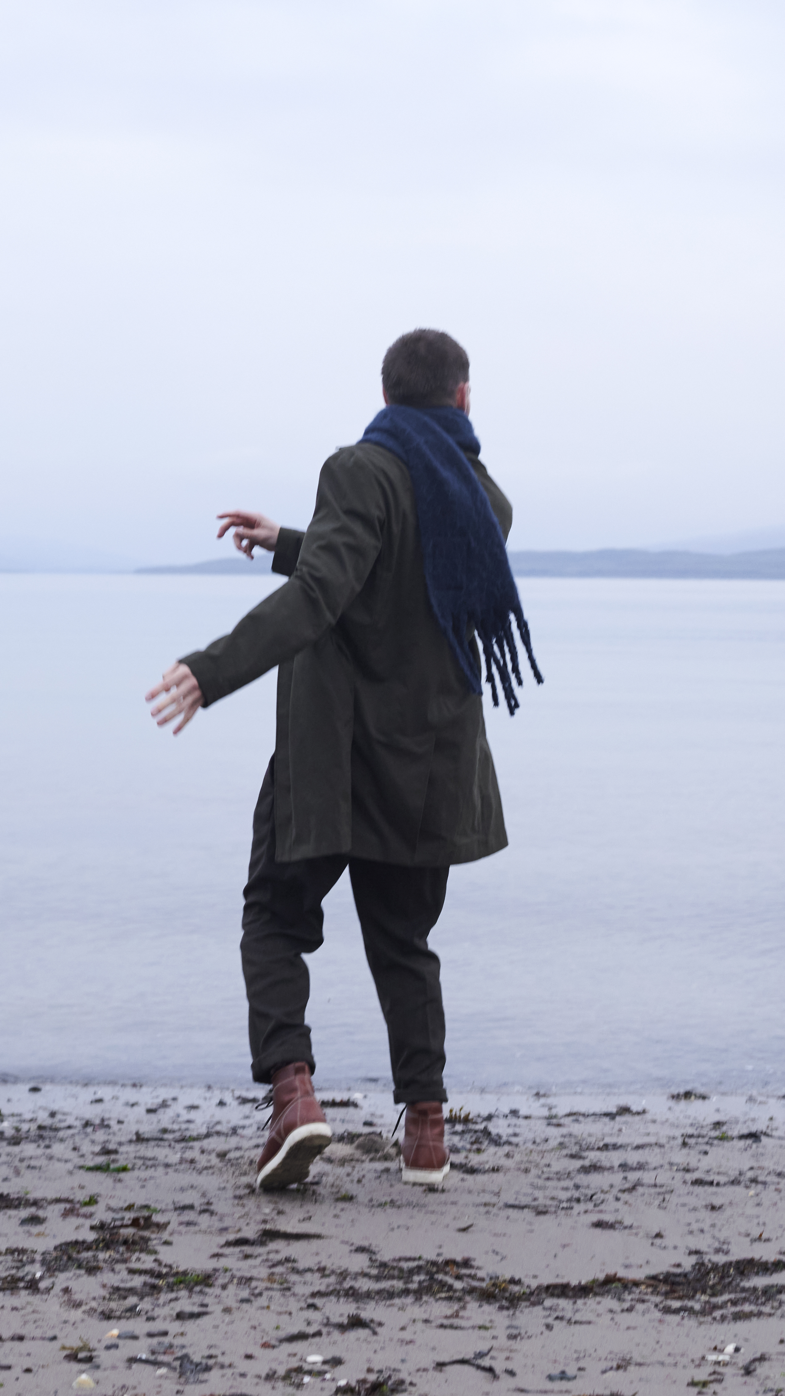 A man skips stones on a sandy shoreline. The sea is still and calm.