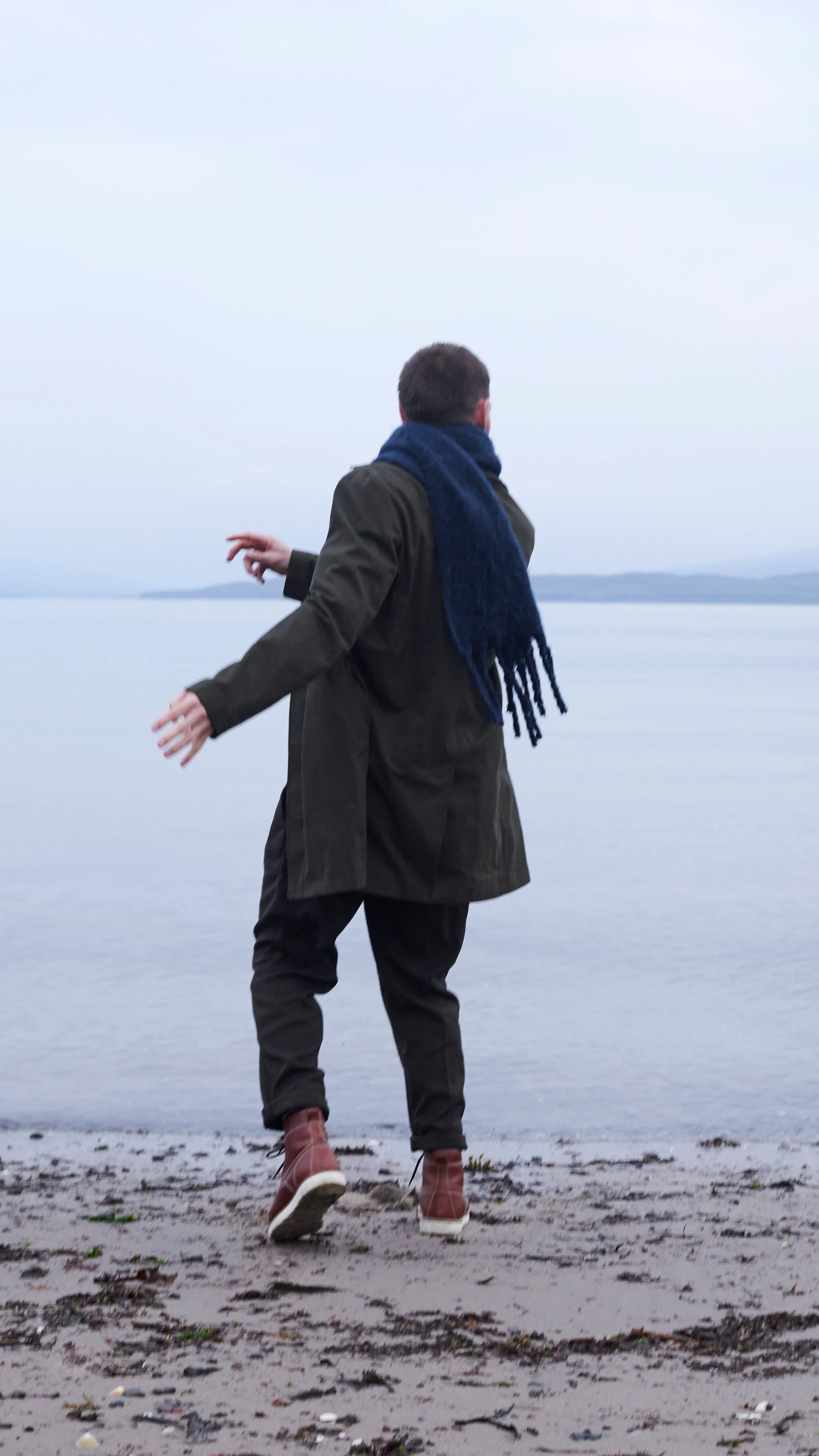 A man skips stones on a sandy shoreline. The sea is still and calm.
