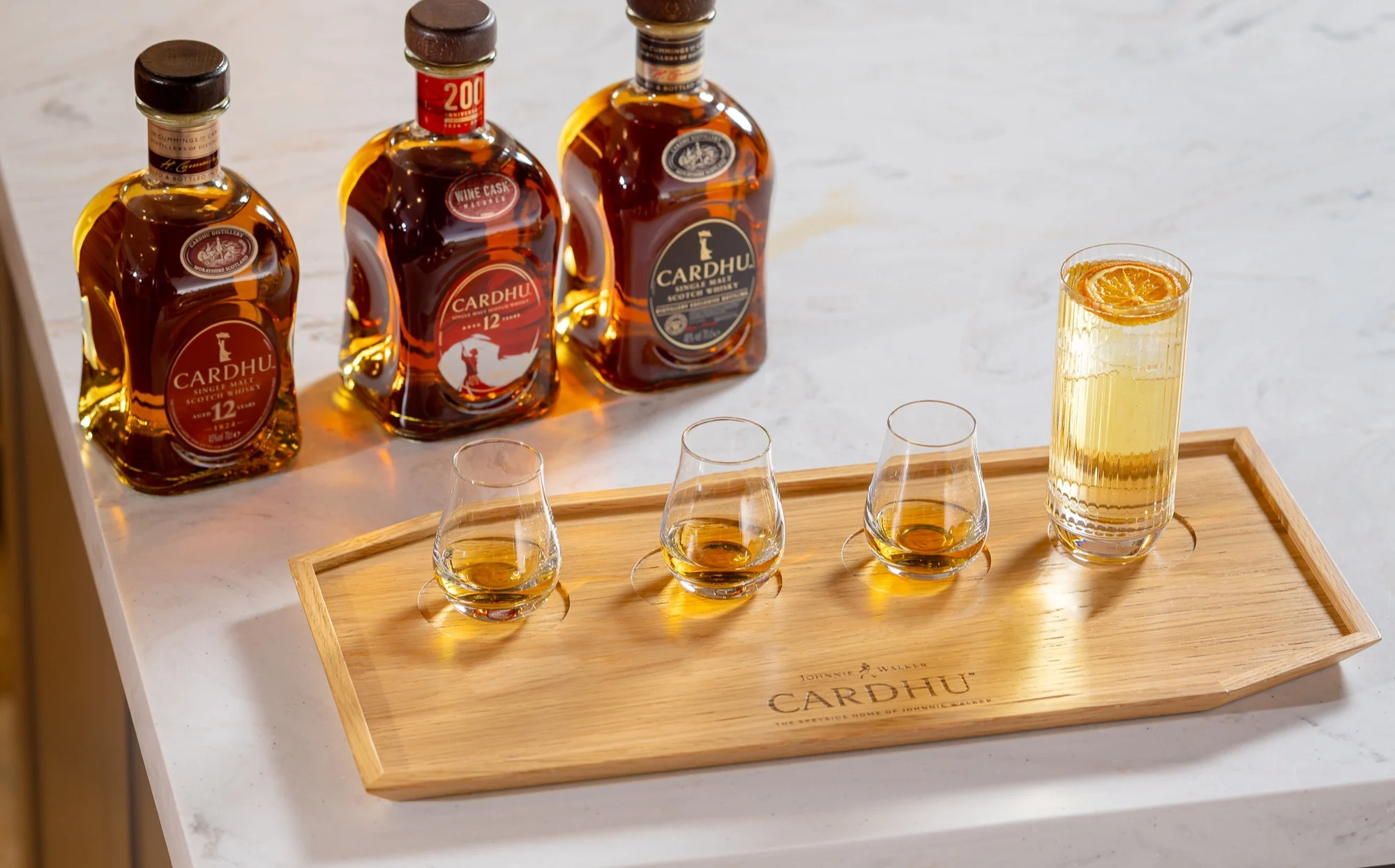 Three bottles of Cardhu whisky sit in a row on a marble countertop. In front sits a wooden tray with the word ‘Cardhu’ carved into it. On the tray sits three dram glasses containing whisky, a whisky cocktail with ice and an orange slice.