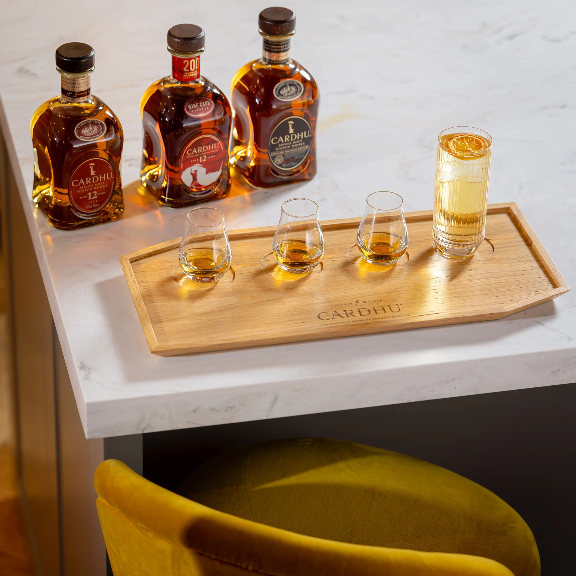 Three bottles of Cardhu whisky sit in a row on a marble countertop. In front sits a wooden tray with the word ‘Cardhu’ carved into it. On the tray sits three dram glasses containing whisky, a whisky cocktail with ice and an orange slice.