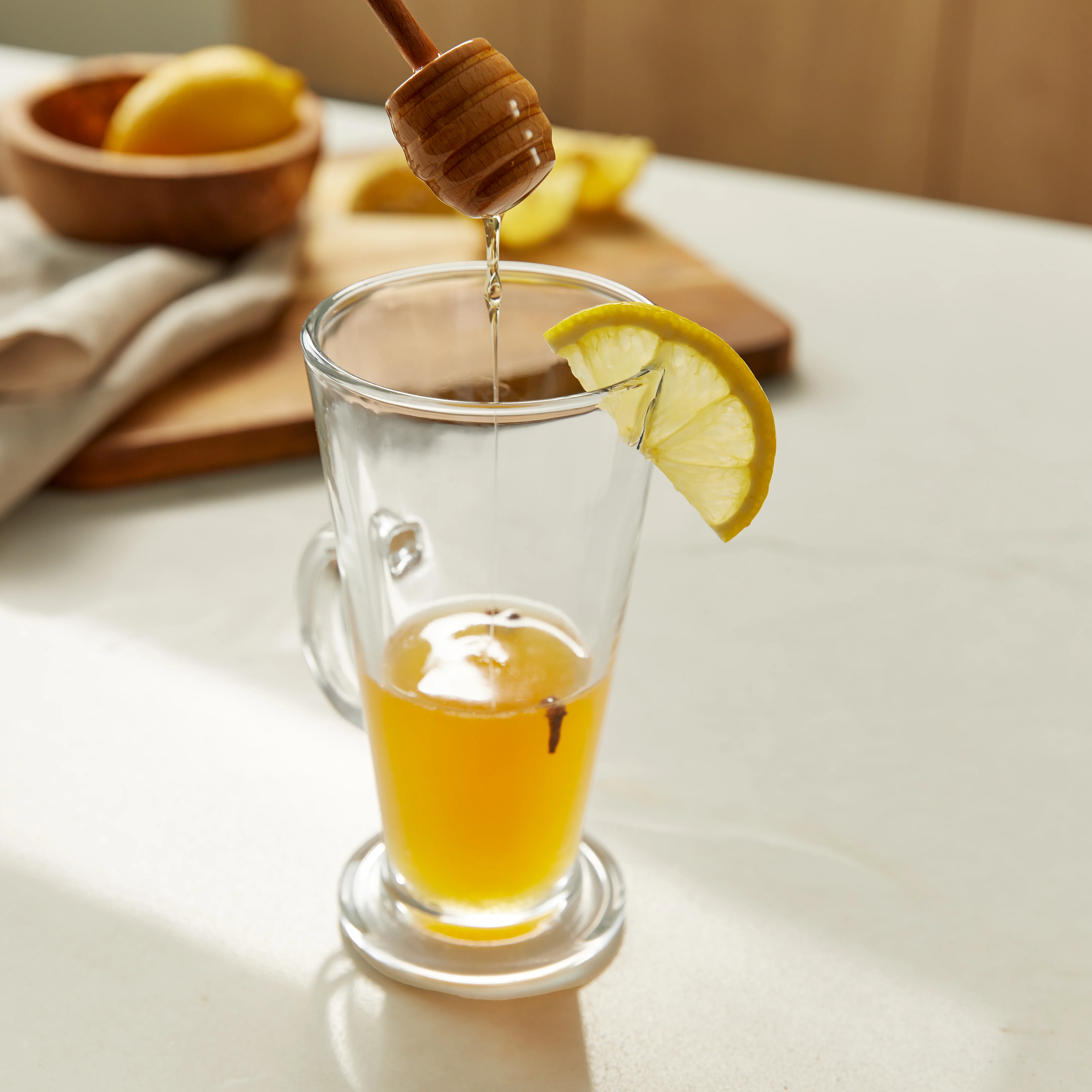 Honey drips off a honey dipper into a glass latte glass containing a light brown liquid, with a lemon slice on the rim