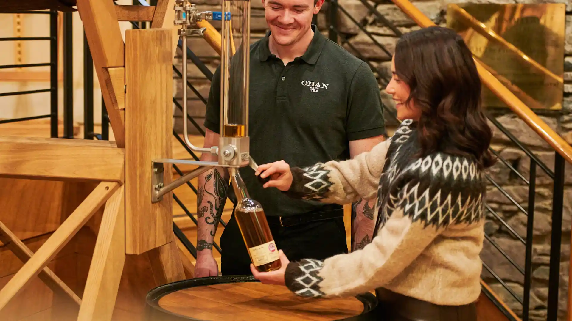 A woman fills a bottle of whisky through a tube, which is the Oban 'bottle your own experience'. An employee of Oban distillery looks on, smiling.